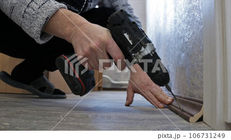 fixing a dark wooden floor plinth with a screwdriver in a man's hand, fixing a brown plinth between a floor with gray linoleum and a wall with light wallpaper using a screwdriver 106731249