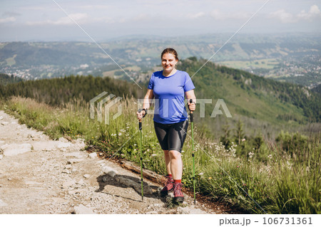 Happy woman hiker with backpack standing on the slope of mountain ridge against mountains. 30s Women holding hiking sticks. Travel and active lifestyle concept, outdoor activities, full body 106731361
