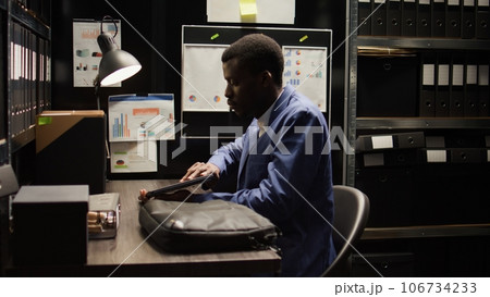 Private investigator, laptop bag in hand, enters office and starts working. African American law officer positions his personal computer on the desk and examines case details related to a suspect. Private investigator, laptop bag in hand, enters office and starts working. African American law officer positions his personal computer on the desk and examines case details related to a suspect. 106734233