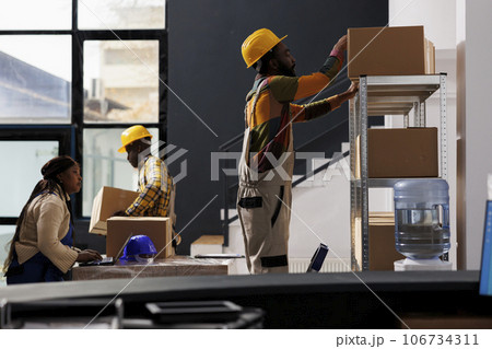 Retail storehouse employees preparing customer purchase for dispatching. All black warehouse order pickers team reading pick ticket on laptop and taking cardboard box from shelf in storage room 106734311