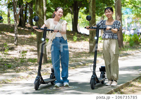 緑が美しい並木道を電動キックボードで爽快に走る若くてかわいい女性2人|電動キックボードイメージ 緑が美しい並木道を電動キックボードで爽快に走る若くてかわいい女性2人|電動キックボードイメージ 106735785