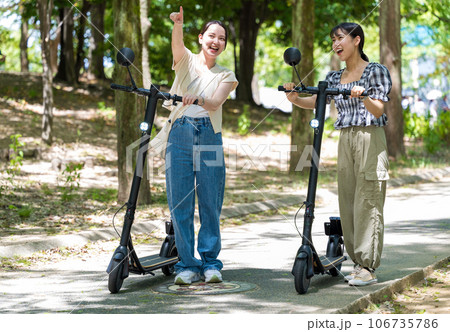 緑が美しい並木道を電動キックボードで爽快に走る若くてかわいい2人の女性｜電動キックボードイメージ 106735786