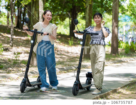 緑が美しい並木道を電動キックボードで移動する若くてかわいい2人の女性｜電動キックボードイメージ 106735787