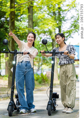 緑が美しい並木道を電動キックボードで移動する若くてかわいい2人の女性|電動キックボードイメージ 緑が美しい並木道を電動キックボードで移動する若くてかわいい2人の女性|電動キックボードイメージ 106735793