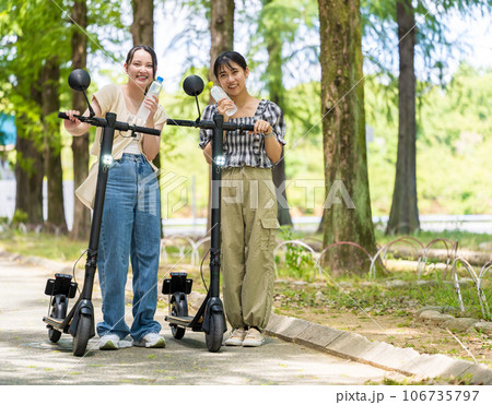 緑が美しい並木道を電動キックボードで散策し、水分補給するかわいい2人の女性｜電動キックボードイメージ 106735797