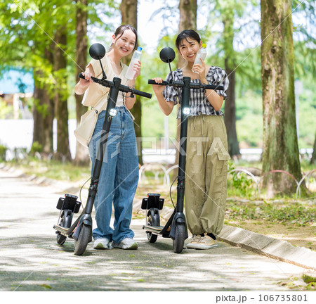 緑が美しい並木道を電動キックボードで散策し、水分補給するかわいい2人の女性|電動キックボードイメージ 緑が美しい並木道を電動キックボードで散策し、水分補給するかわいい2人の女性|電動キックボードイメージ 106735801
