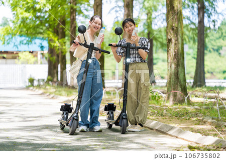 緑が美しい並木道を電動キックボードで散策し、水分補給するかわいい2人の女性｜電動キックボードイメージ 106735802