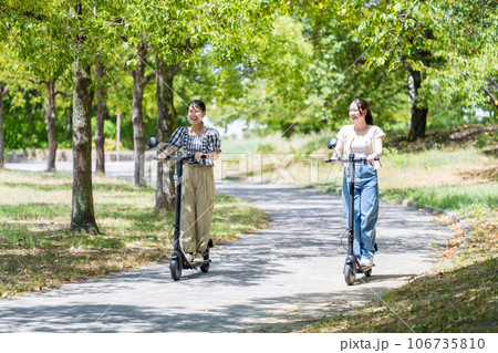 緑が美しい並木道を電動キックボードで走る若くてかわいい2人の女性|電動キックボードイメージ 緑が美しい並木道を電動キックボードで走る若くてかわいい2人の女性|電動キックボードイメージ 106735810