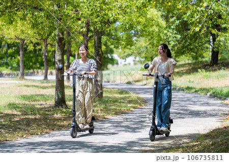 緑が美しい並木道を電動キックボードで走る若くてかわいい2人の女性|電動キックボードイメージ 緑が美しい並木道を電動キックボードで走る若くてかわいい2人の女性|電動キックボードイメージ 106735811