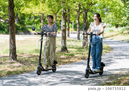 緑が美しい並木道を電動キックボードで走る若くてかわいい2人の女性|電動キックボードイメージ 緑が美しい並木道を電動キックボードで走る若くてかわいい2人の女性|電動キックボードイメージ 106735812