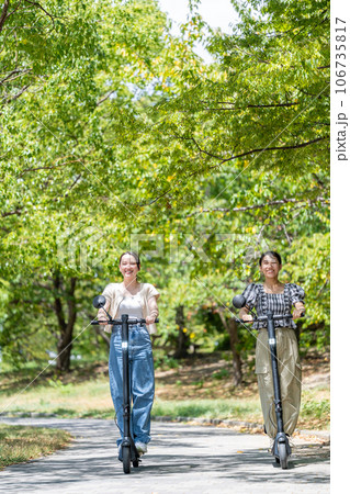 緑が美しい並木道を電動キックボードで走る若くてかわいい2人の女性｜電動キックボードイメージ 106735817