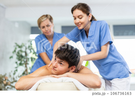 Female professional physiotherapist helps girl student to perform back massage to male client. Female professional physiotherapist helps girl student to perform back massage to male client. 106736163