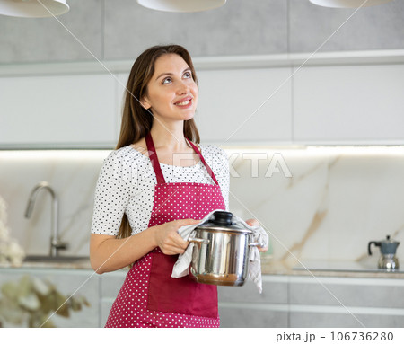 Smiling girl is standing in kitchen in dotted apron and holding stainless steel pot with lid 106736280