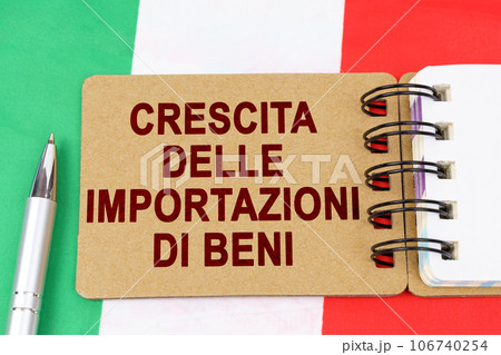 Economy and finance concept. On the flag of Italy lies a pen and a notebook with the inscription - growth in imports of goods. Text in Italian. Economy and finance concept. On the flag of Italy lies a pen and a notebook with the inscription - growth in imports of goods. Text in Italian. 106740254