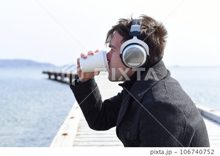 Young man listening to music and drinking coffee, sitting alone on pier on a sunny day 106740752