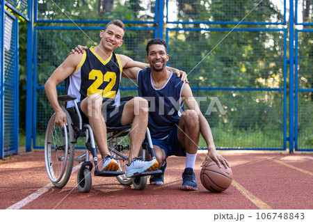 Man playing basketball with disabled friend in wheelchair at outdoor court. 106748843