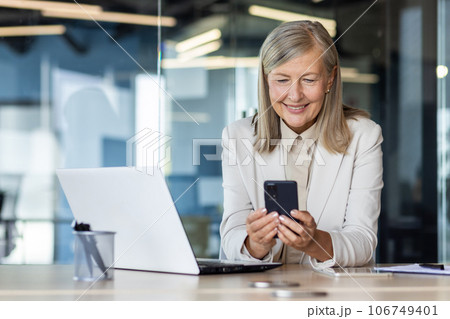 Senior mature experienced woman at workplace inside office, business woman happy smiling using phone sitting with laptop, gray haired woman boss using app on smartphone typing message. Senior mature experienced woman at workplace inside office, business woman happy smiling using phone sitting with laptop, gray haired woman boss using app on smartphone typing message. 106749401