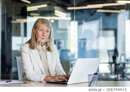 Portrait of serious mature thinking businesswoman, gray haired female boss looking at camera, at workplace inside office, financier with laptop, confident accountant investor. 106749413
