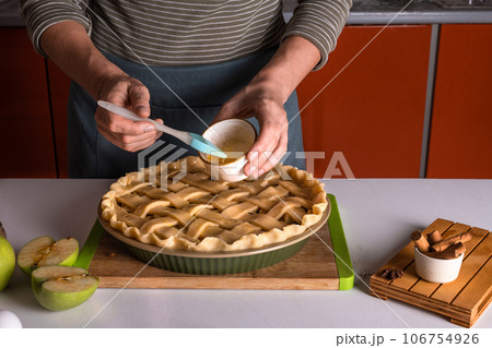 Woman spreading eggs on the raw apple pie with a cooking brush in a bakery dish, recipe Woman spreading eggs on the raw apple pie with a cooking brush in a bakery dish, recipe 106754926