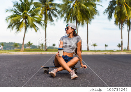 Young beautiful woman in a cap sitting on a skateboard and drinking coffee 106755243