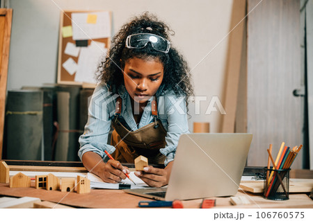 Carpenter america black woman curly hair sketch making notes in work paper while standing at desk with laptop computer in wood workshop, young female working learning online at woodshop Carpenter america black woman curly hair sketch making notes in work paper while standing at desk with laptop computer in wood workshop, young female working learning online at woodshop 106760575