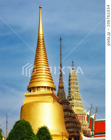 A temple in Thailand with golden and white spires. The temple has a large golden spire. The sky is blue and the temple is bathed in sunlight. 106762834