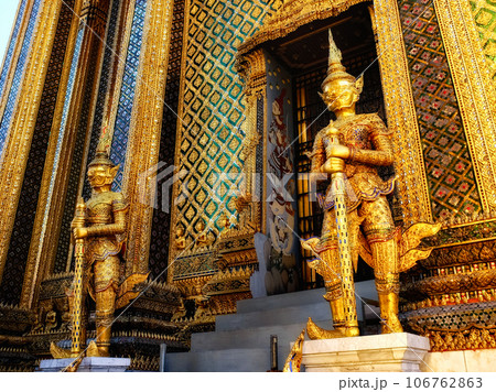 Two golden statues of demon holding weapons stand guard in front of an ornately decorated temple with gold walls. 106762863