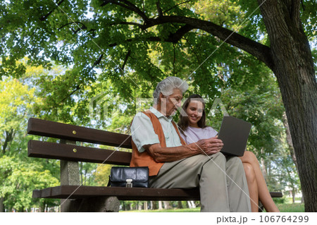 The grandfather and granddaughter are looking at photos on notebook, sitting on a bench in a city park. 106764299