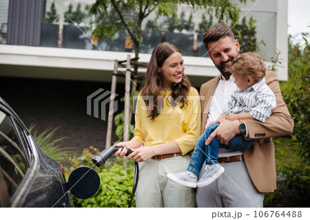 Happy family standing beside their car and charging electric car on the street. 106764488
