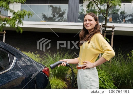 Close up of beautiful woman plugging charger in her electric car. Progressive woman charging her electric car on the street. Close up of beautiful woman plugging charger in her electric car. Progressive woman charging her electric car on the street. 106764491