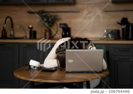 A young beautiful brunette woman is sleeping on a table in the kitchen, tired of working on a laptop A young beautiful brunette woman is sleeping on a table in the kitchen, tired of working on a laptop 106764496