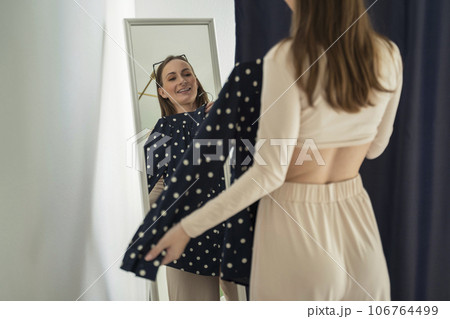 Pretty young woman standing and choosing clothes, trying on a shirt, a woman getting dressed, getting ready before leaving for work, looking at her reflection in the mirror 106764499