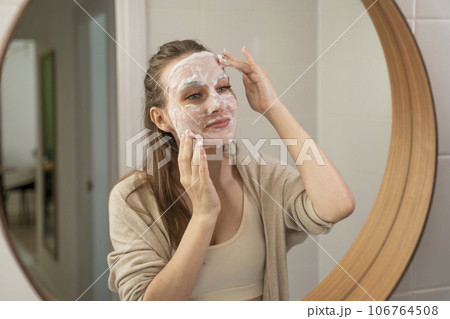 A young woman using an emollient facial moisturizer for facial skin care. Cosmetic procedures. A woman puts makeup on her face while relaxing in the bathroom A young woman using an emollient facial moisturizer for facial skin care. Cosmetic procedures. A woman puts makeup on her face while relaxing in the bathroom 106764508