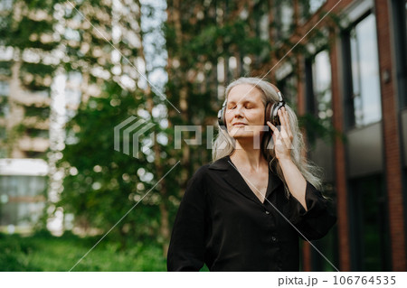 Portrait of a beautiful woman with gray hair listening to music through wireless headphones in city park. 106764535