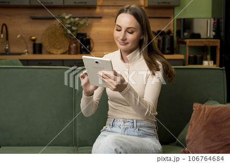 Smiling brunette woman at home is sitting on a green sofa and using a digital tablet Smiling brunette woman at home is sitting on a green sofa and using a digital tablet 106764684