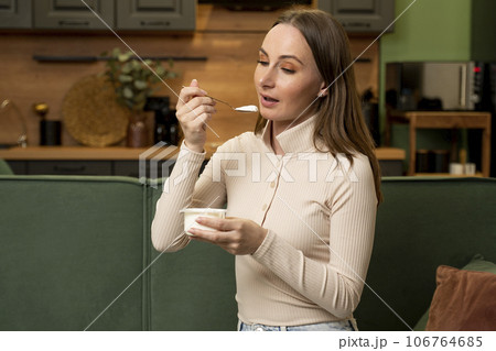 Happy brunette woman eats delicious yogurt at home sitting on a green sofa Happy brunette woman eats delicious yogurt at home sitting on a green sofa 106764685
