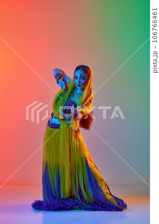 Young beautiful woman in traditional indian dress and makeup dancing against gradient studio background in neon light 106766461