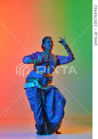 Odissi dance. Beautiful mature indian woman in traditional dress dancing indian dance against gradient studio background in neon light Odissi dance. Beautiful mature indian woman in traditional dress dancing indian dance against gradient studio background in neon light 106766482