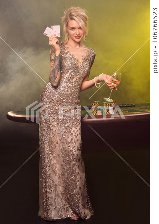 Blonde woman with a beautiful hairstyle and perfect make-up is posing with two playing cards in her hands. Casino, poker. Blonde woman with a beautiful hairstyle and perfect make-up is posing with two playing cards in her hands. Casino, poker. 106766523