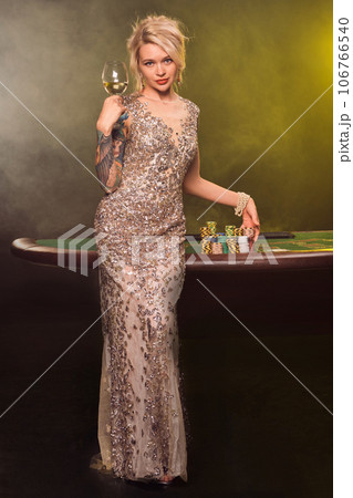 Blonde woman with a beautiful hairstyle and perfect make-up is posing with a glass of champagne in her hand. Casino, poker. Blonde woman with a beautiful hairstyle and perfect make-up is posing with a glass of champagne in her hand. Casino, poker. 106766540