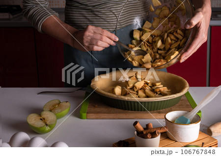 Woman pouring spiced apples stuffing for the apple pie in the bakery dish with dough Woman pouring spiced apples stuffing for the apple pie in the bakery dish with dough 106767848
