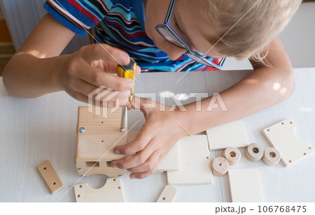 An 8-year-old boy assembles a wooden constructor by himself with a screwdriver. Close-up hands. An 8-year-old boy assembles a wooden constructor by himself with a screwdriver. Close-up hands. 106768475