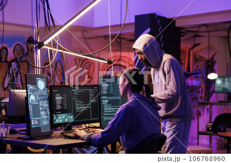 Hackers coding malicious software on computer together while working in abandoned warehouse. Internet thieves in hoods planning phishing attack and programming malware in dark room 106768960