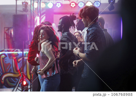 Young woman making passionate dance moves while clubbing with friends at discotheque. Crowd partying on vibrant dancefloor while attending disco party with live music in nightclub Young woman making passionate dance moves while clubbing with friends at discotheque. Crowd partying on vibrant dancefloor while attending disco party with live music in nightclub 106768988