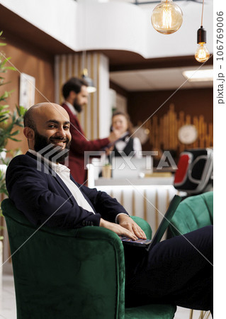 Guest prepares for business meeting, working on laptop in lounge area lobby before room registration. Businessman engaging in productive work to kill time while he waits for conference. Guest prepares for business meeting, working on laptop in lounge area lobby before room registration. Businessman engaging in productive work to kill time while he waits for conference. 106769006