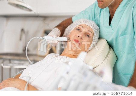 Elderly female client receiving hardware ultrasonic to revitalize and tighten facial skin at cosmetology clinic Elderly female client receiving hardware ultrasonic to revitalize and tighten facial skin at cosmetology clinic 106771014