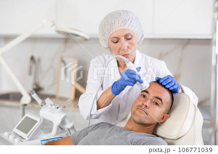 Female cosmetologist performing anti-aging face phototherapy procedure for male client in clinic of aesthetic clinic 106771039