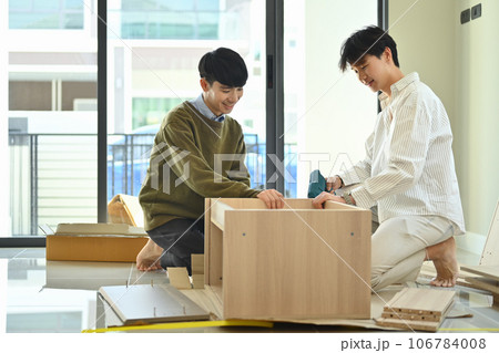 Cheerful young gay male couple using electric drill tool to assembling furniture in their new home 106784008