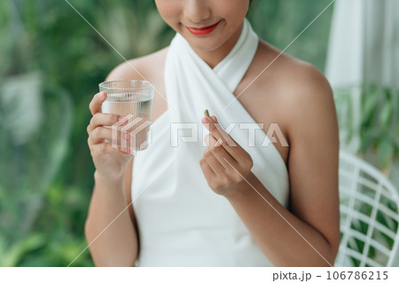 Young woman at home near window holding pill and glass of fresh water 106786215