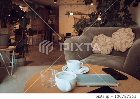 Coffee cups and book on wooden table. Armchair with cushions and table with coffee cups in cafe. Breakfast table in restaurant. Modern urban lifestyle. Morning drinks. Coffee break.  106787553